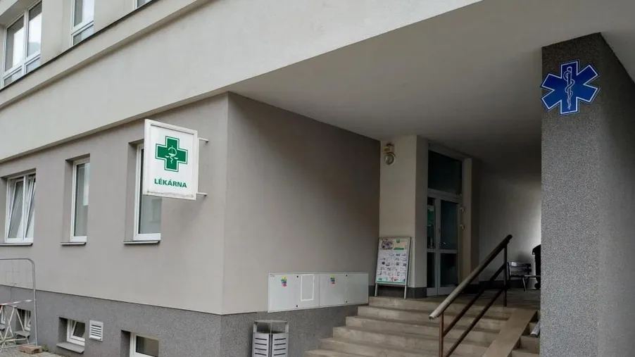 Vchod do lékárny v přízemí se zeleným lékárenským křížem a modrým znakem hvězdy života. Přístup po několika schodech s kovovým zábradlím.